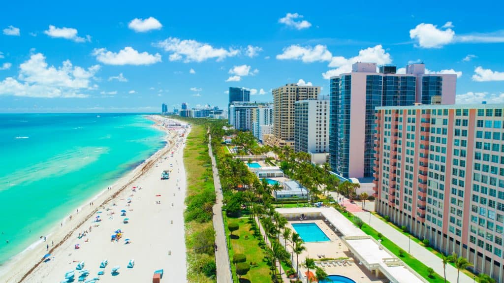 Photo is an aerial view of South Beach located in Miami that features large condo buildings, white sand, and blue ocean water.