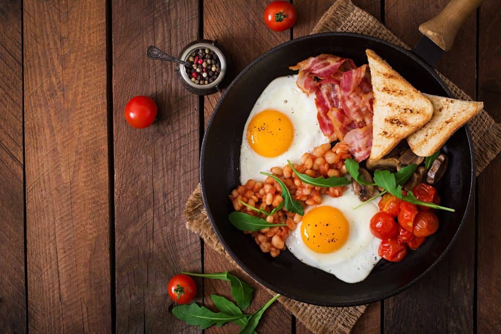 Over easy eggs, with bacon and toast with roasted tomatoes and baked beans, set served in a skillet.