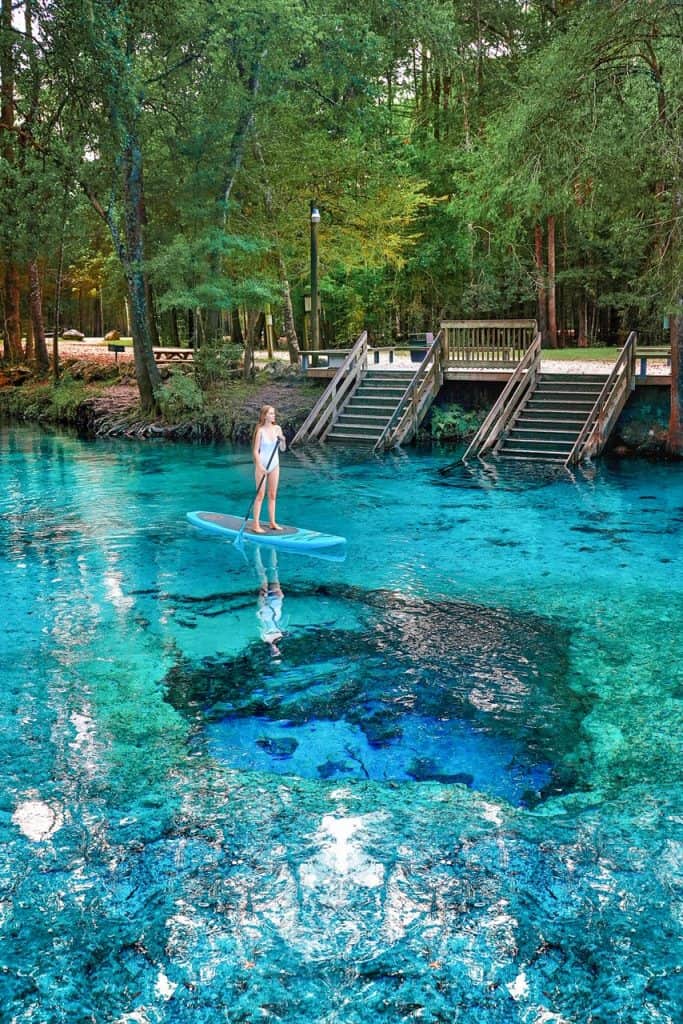 woman paddle boarding over devils eye spring at ginnie springs in florida. the water is blue and she is wearing a white swimsuit