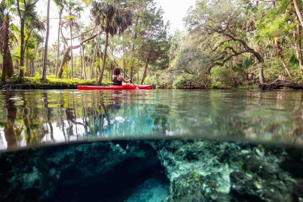A kayaker paddles on Seven Sister Springs; the camera also captures the view underwater, which is crystal clear, revealing rock formations below.