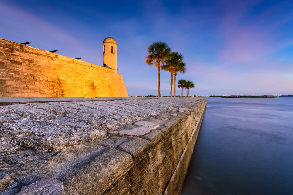 A small, stone Florida castle at sunset next to the water, with palm trees beside it