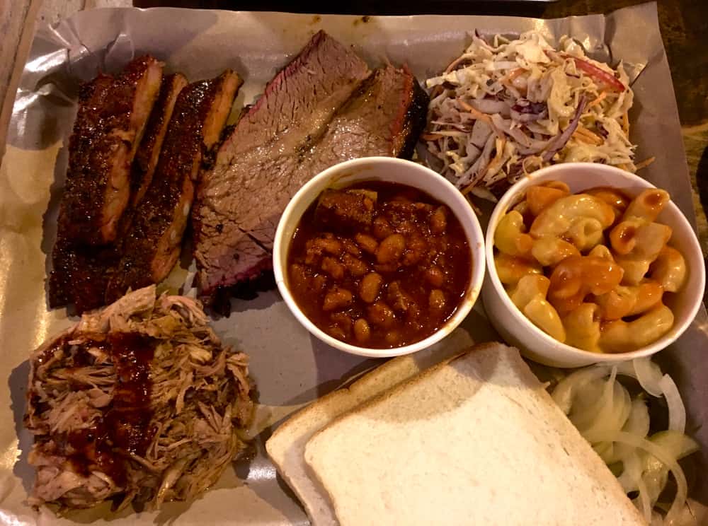 A plate of bbq food with pulled pork, beans, mac and cheese, slaw and brisket with slices of white bread