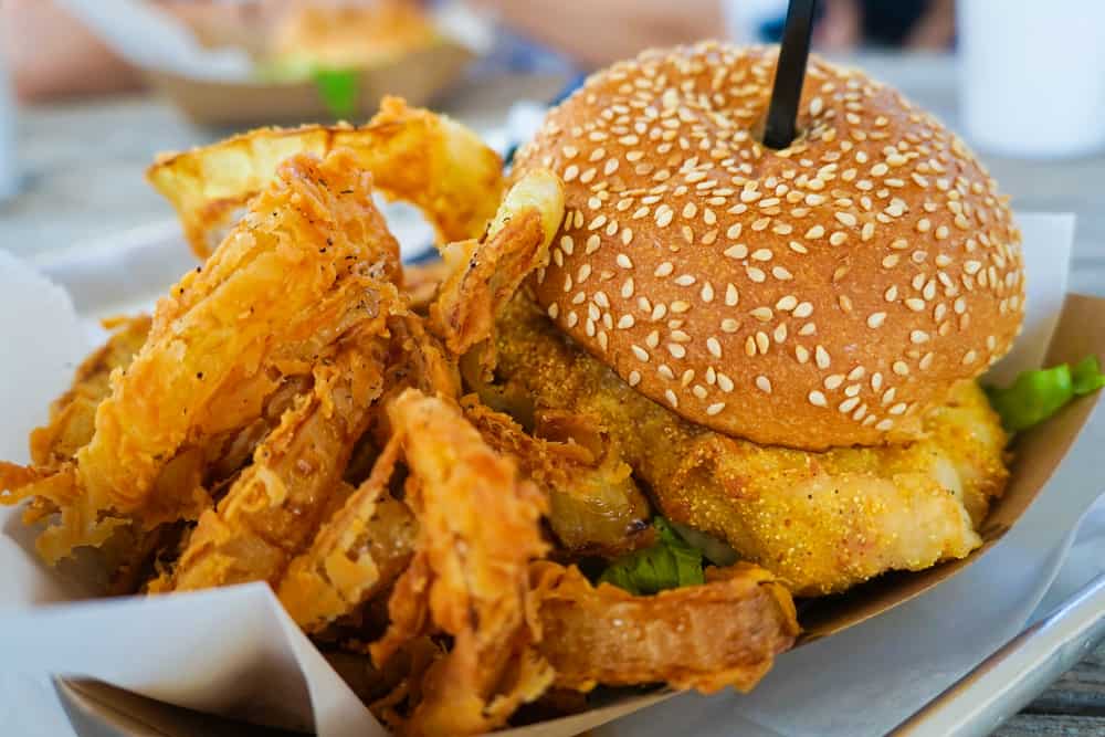 a fried fish sandwich on sesame bun with fries and onion rings