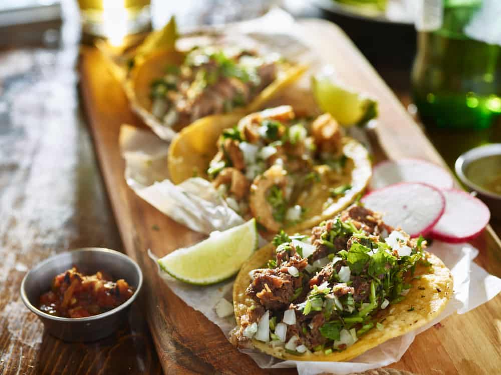 A plate of Mexican style tacos with meat cilantro and hard shell served with limes on a wood tray
