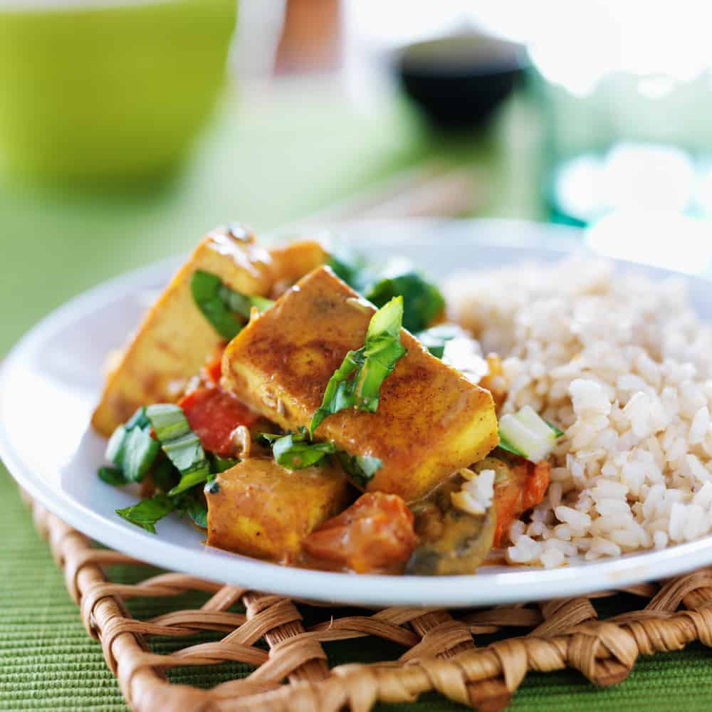 A plate of tofu in a curry sauce served with rice over a bamboo placemat