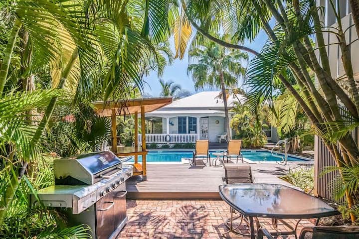 View of a bbq, pool and benches on shaded patio, surrounded by palm trees.