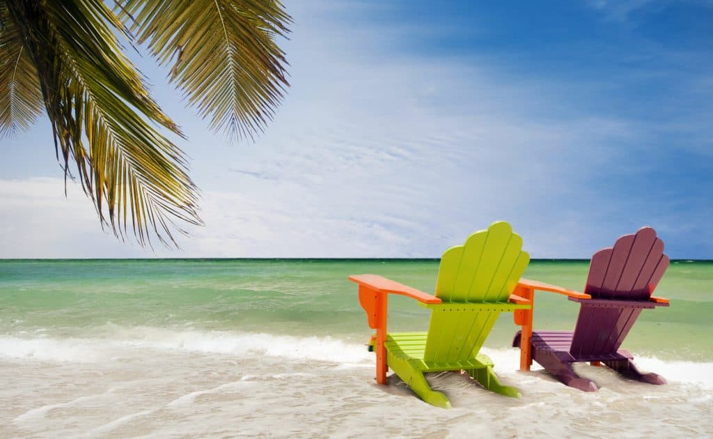 two colorful chairs sitting in the water on a beach under a palm tree