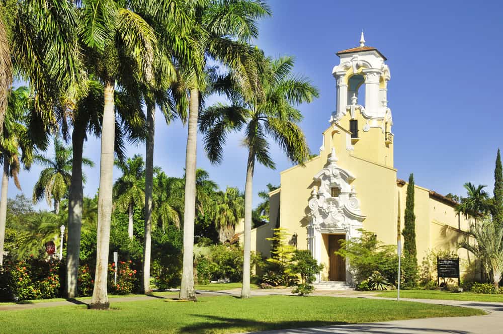 The yellow and white Coral Gables Congregational Church with palm trees in front.
