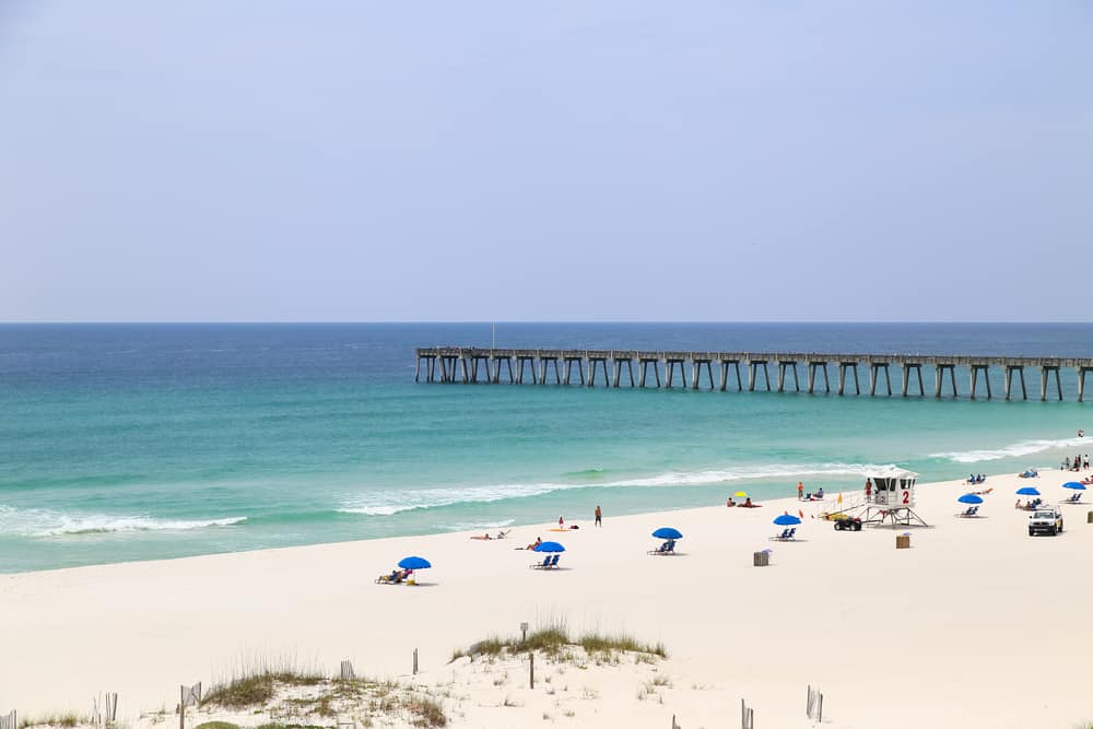 A pier jutting out into the waters of Pensacola Beach, where people on weekend getaways in Florida sit under blue umbrellas.