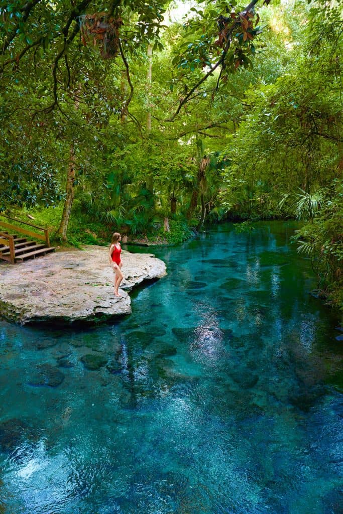 A woman in red swim suit standing on edge of bright blue spring surrounded by greenery during weekend getaways in Florida.