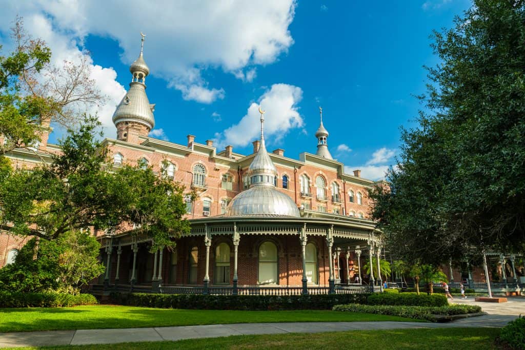 The stunning architecture of the Henry B. Plant Museum. There is a lawn, trees, and gardens surrounding the brick building. 