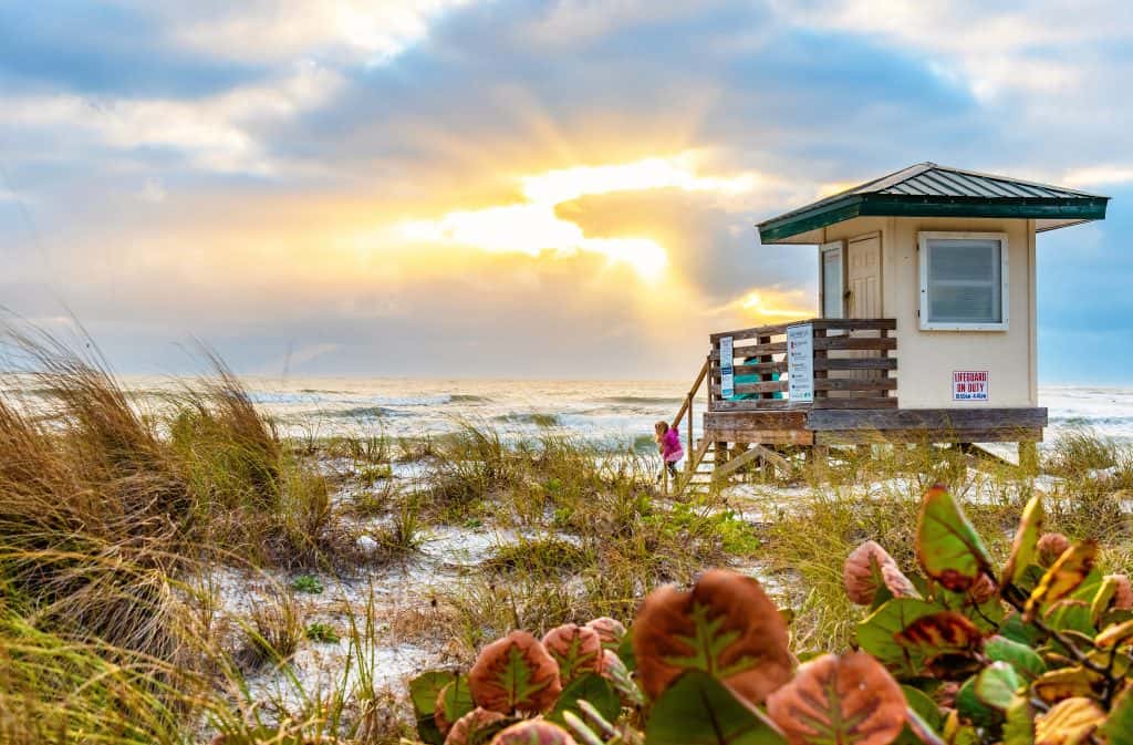 A lifeguard tower stands among the reeds in the sands of Lido Key Beach, one of the best things to do in Sarasota, as the sun peeks through clouds and waves crash on the shore.