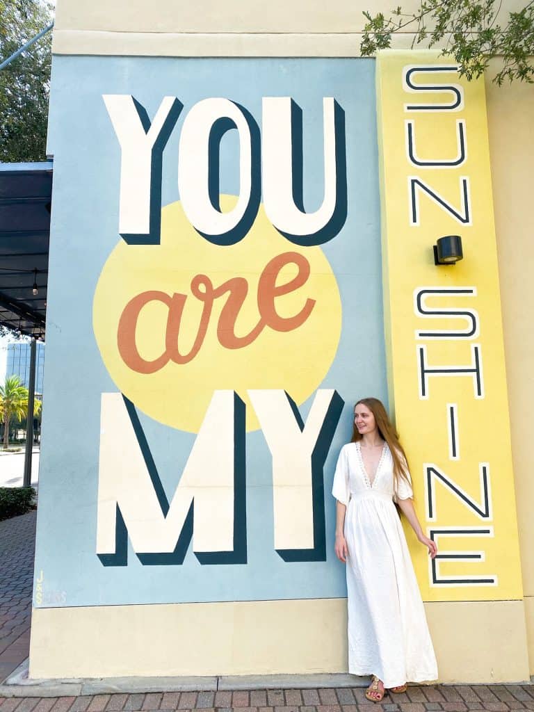 A woman in a long dress stands in front of a "you are my sunshine" mural, a must for things to do in St. Petersburg, Florida. 