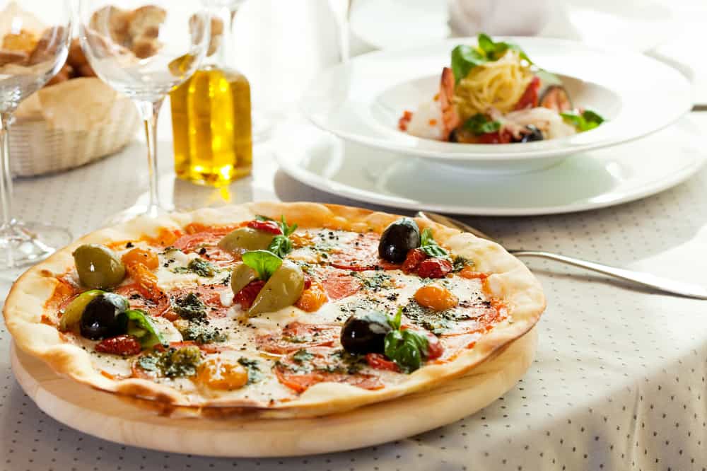 A pizza with olives in the foreground and pasta dish in the background showing the cuisine at restaurants in Naples.