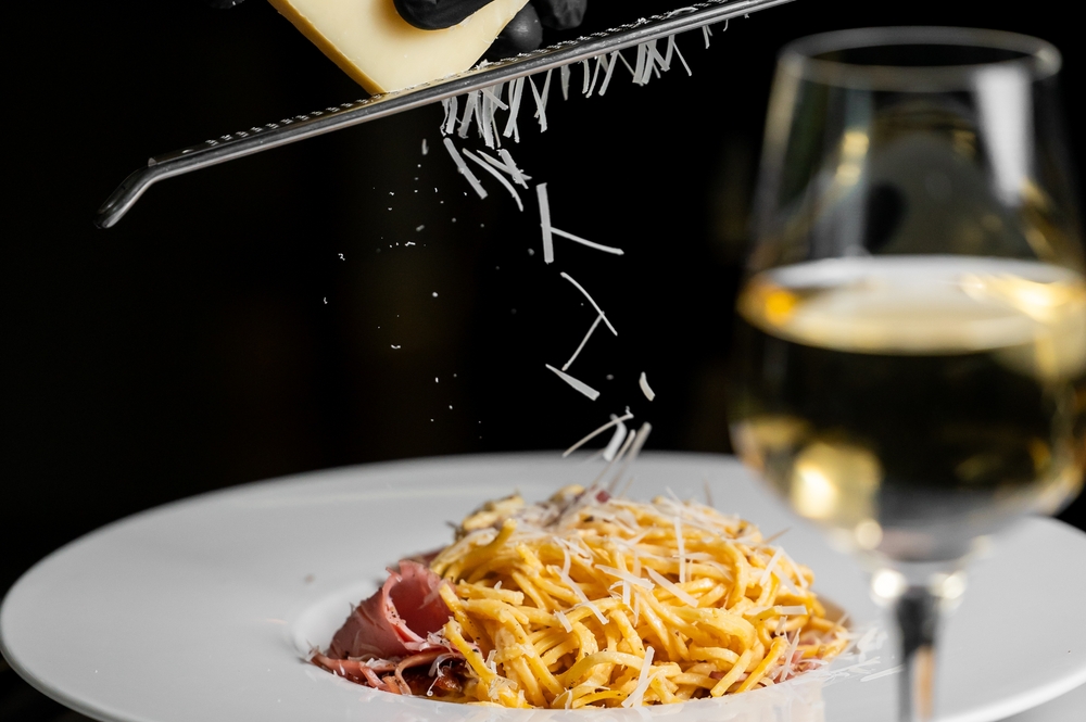 Freshly grated cheese falls onto a plate of pasta with prosciutto, accompanied by a glass of white wine. The close-up highlights the elegance and culinary detail of Italian cuisine.