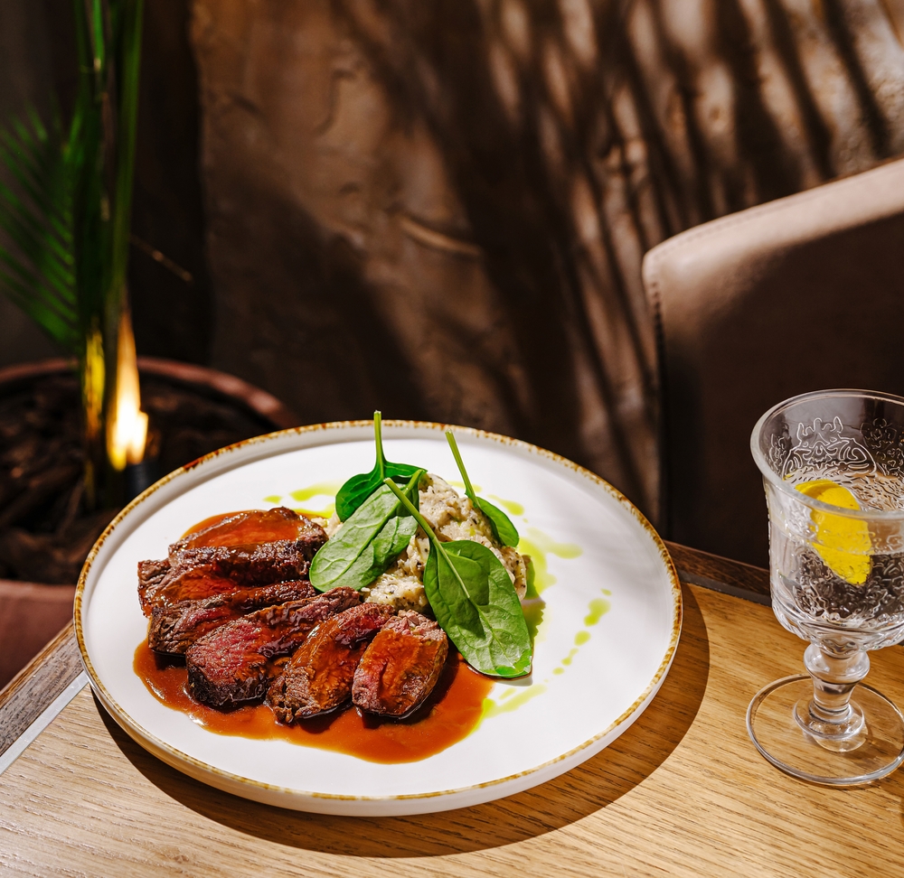 Savor grilled steak slices garnished with fresh spinach and paired with creamy sauce on a white plate. A refreshing drink sits beside the meal in a stylish setting during dinner.