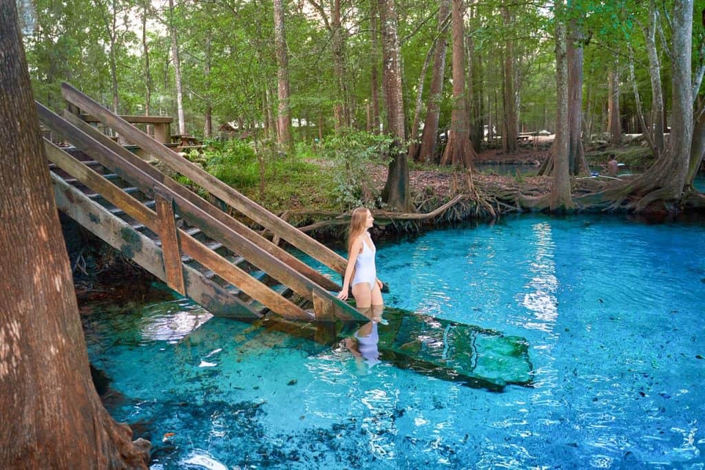 A woman walking down stairs into bright blue water surrounded by trees.