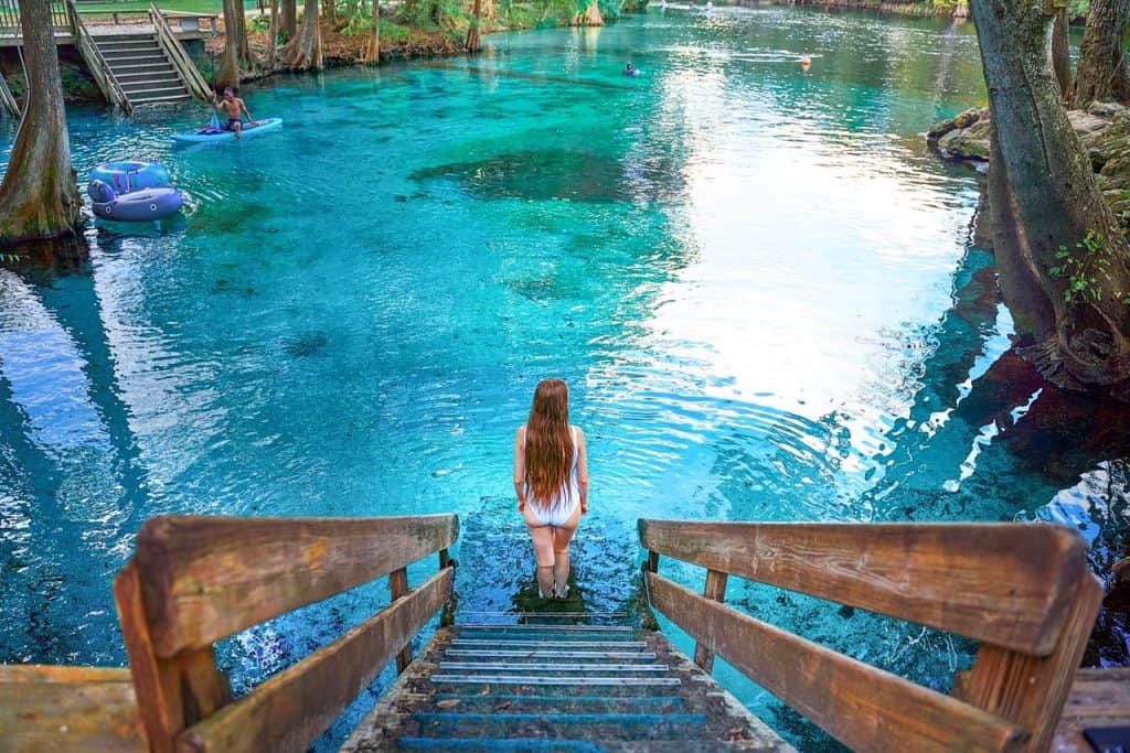 A woman with long hair walking down stairs into bright blue water surrounded by trees.