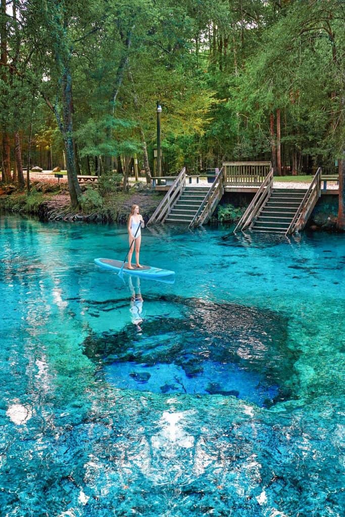 A woman paddle boarding over Devil's Eye Spring at Ginnie Springs in Florida. The water is blue and she is wearing a white swimsuit.