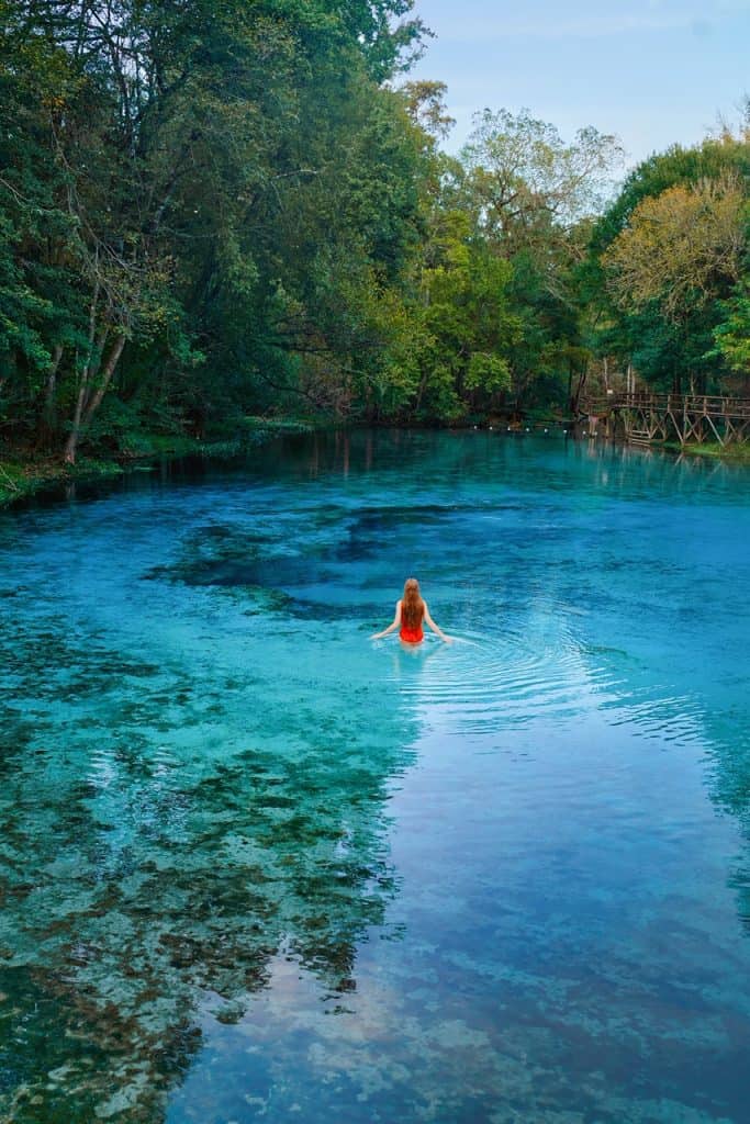 A woman in red bathing suit wading in blue water at Gilchrist Blue Springs in Florida with green trees.