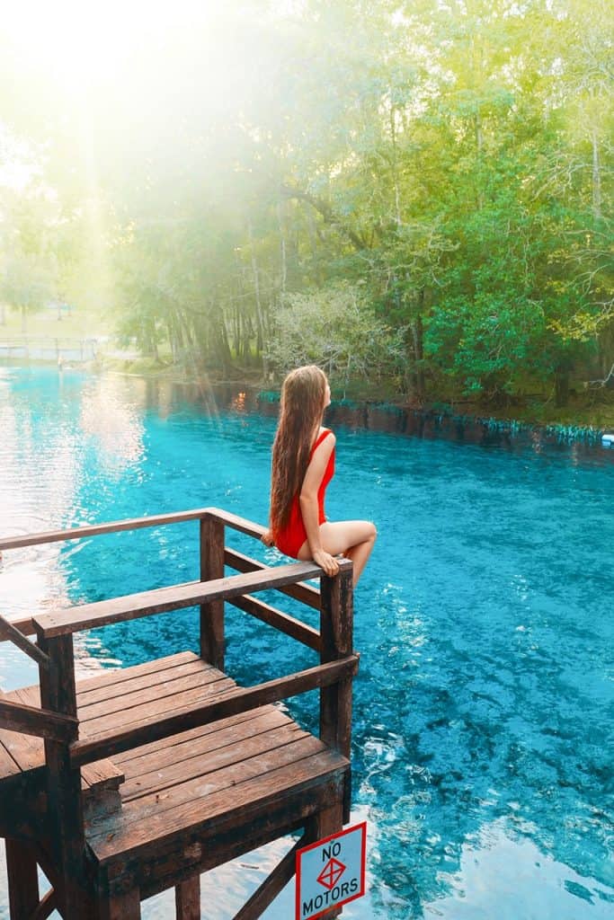 A woman in a red swim suit sits on a wooden railing over Gilchrist Blue Springs.