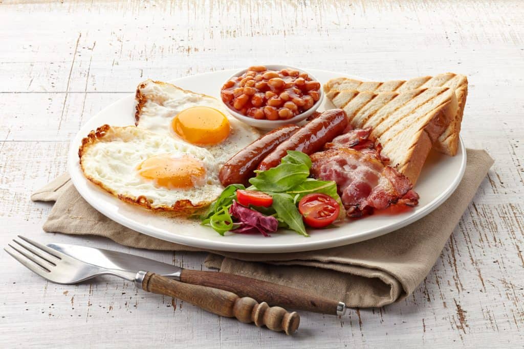 A breakfast of two eggs, beans, toast, sausage, and bacon sit on a plate, ready to be eaten.