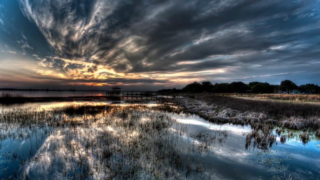 The sun sets over Lake Tohopekaliga, the water reflecting the clouds stretching above as orange and yellow light peeks through. 