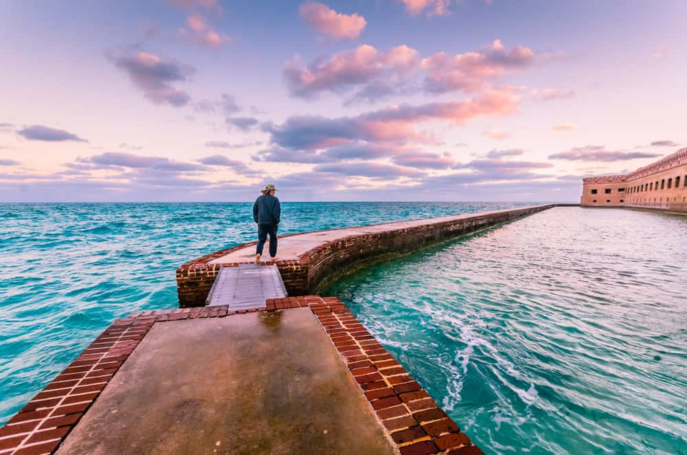 Man walking the moat wall around Fort Jefferson with water on both sides and a sunrise sky overhead.