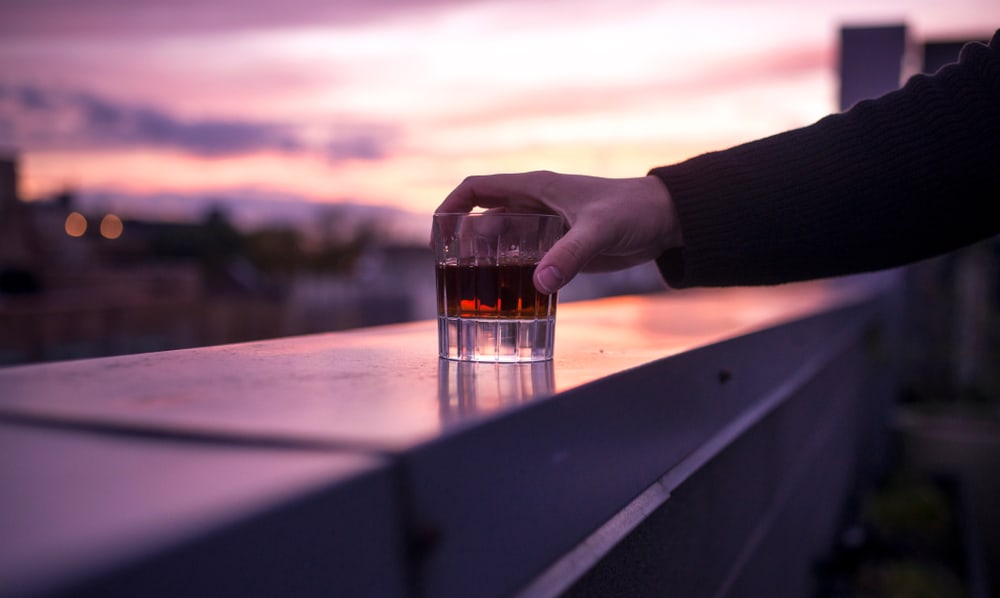 A man's hand holds a whisky on a rooftop bar at sunset.