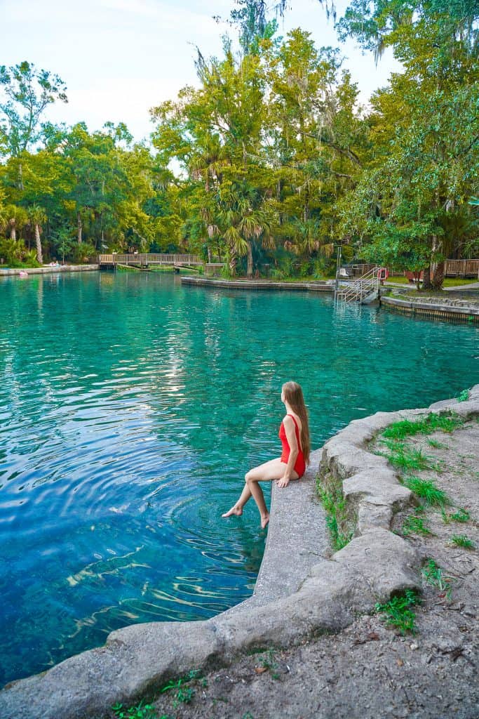 A woman in a red swimsuit sits dips her toes in the blue water of Wekiwa Springs in Central Florida.