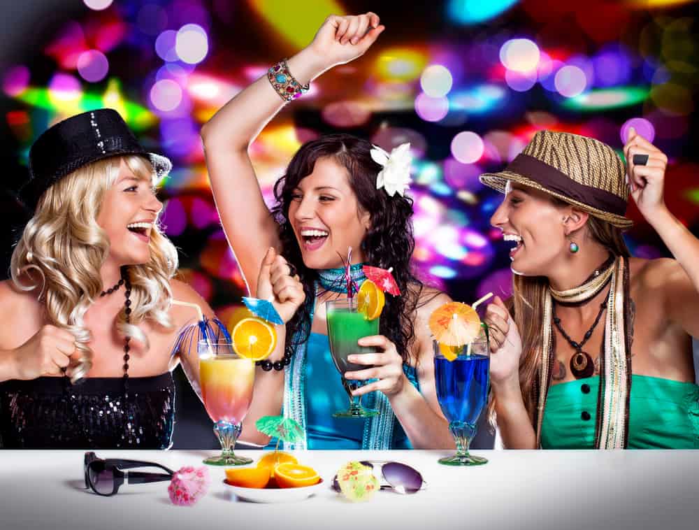 Three girls drinking cocktails in a night club,. Two girls have hats on and one has a flower in her hair. At a Florida bachelorette party