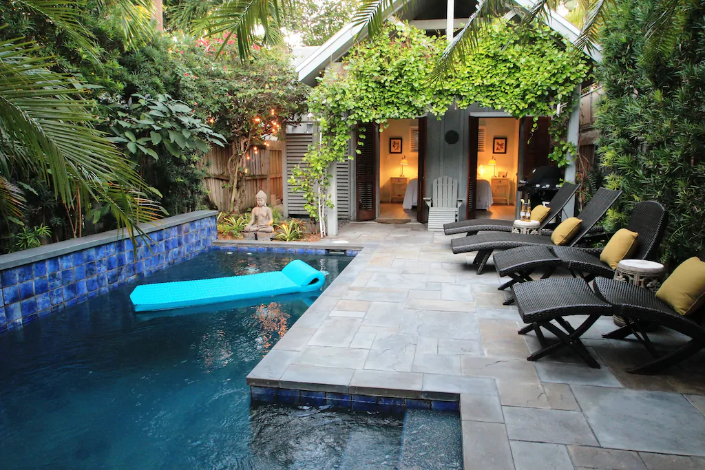 Photo of a pool and lounge area with lush garden surrounding and a cottage. 