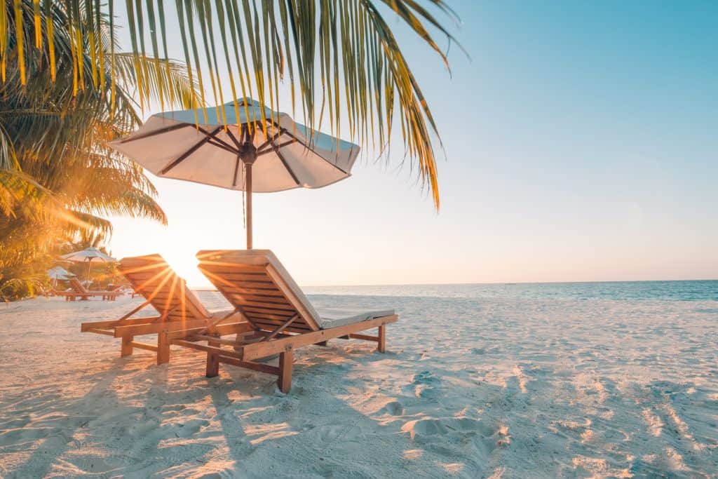 Beach chairs sit on the sand under an umbrella at sunset.