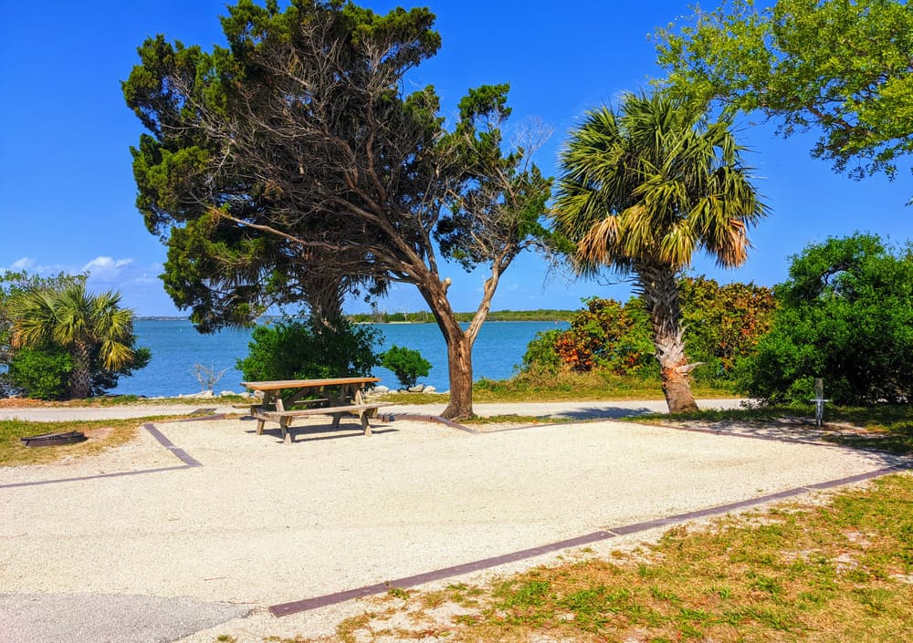 AN empty waterfront gravel camping site with picnic table