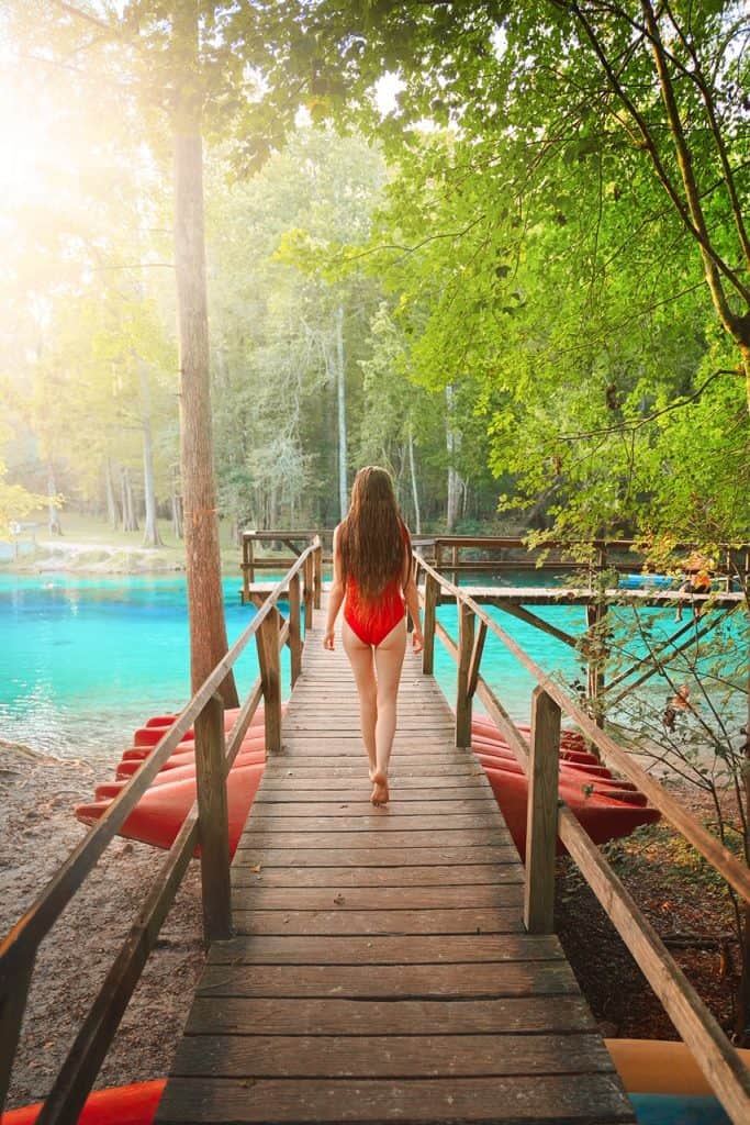 Girl in a red swimsuit walking down a wooden boardwalk at Gilchrist springs with red canoes underneath dock and turquoise water.