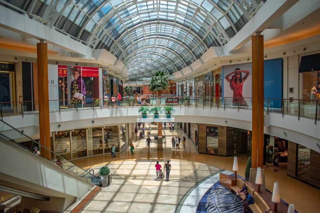 The two-story interior of a mall with guests browsing storefronts under the glass ceiling.