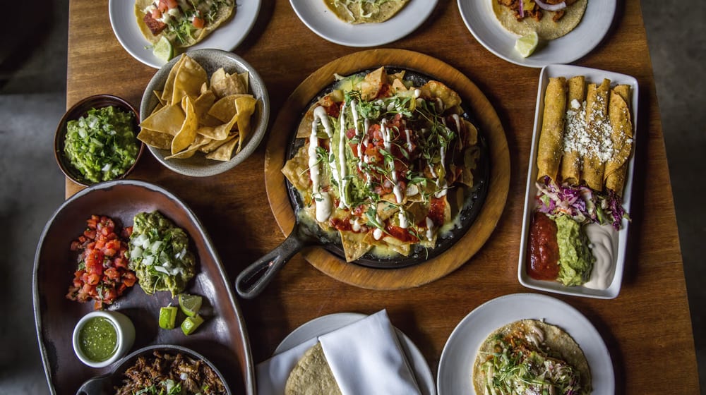 A table full of Mexican dishes, such as loaded nachos, table made guacamole, flaurtas, tacos, and fajitas