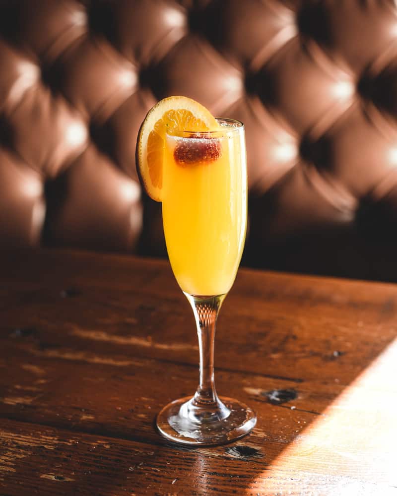 A champagne flute full of mimosa cocktail sits on a wooden surface with a leather booth in the background.