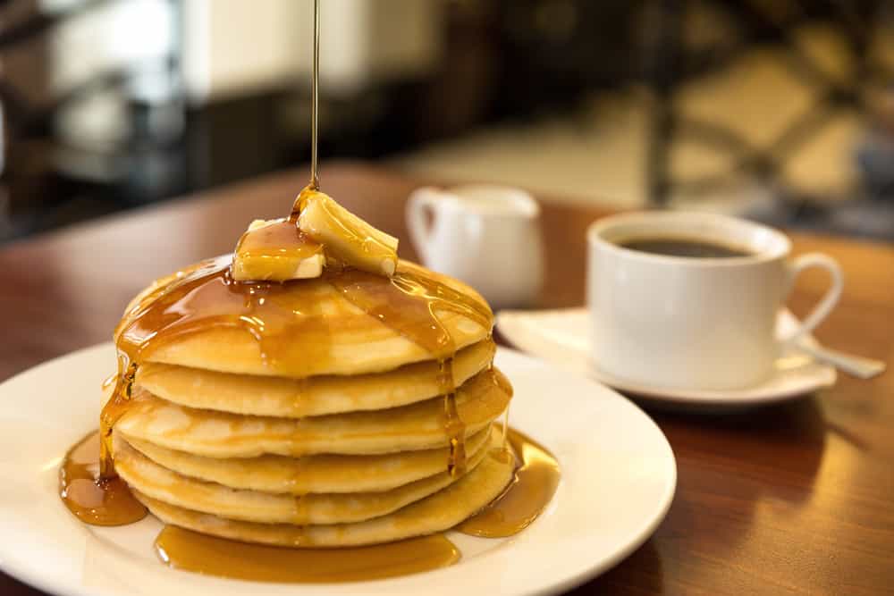 A stack on pancakes topped with butter and syrup sits on a plate, next to a cup of coffee.