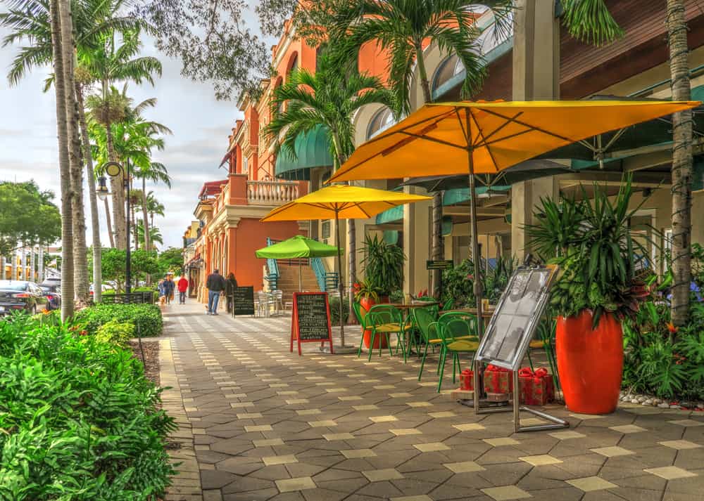 Umbrellas cover chairs and tables for outdoor dining along a palm tree-covered sidewalk in Naples, Florida.