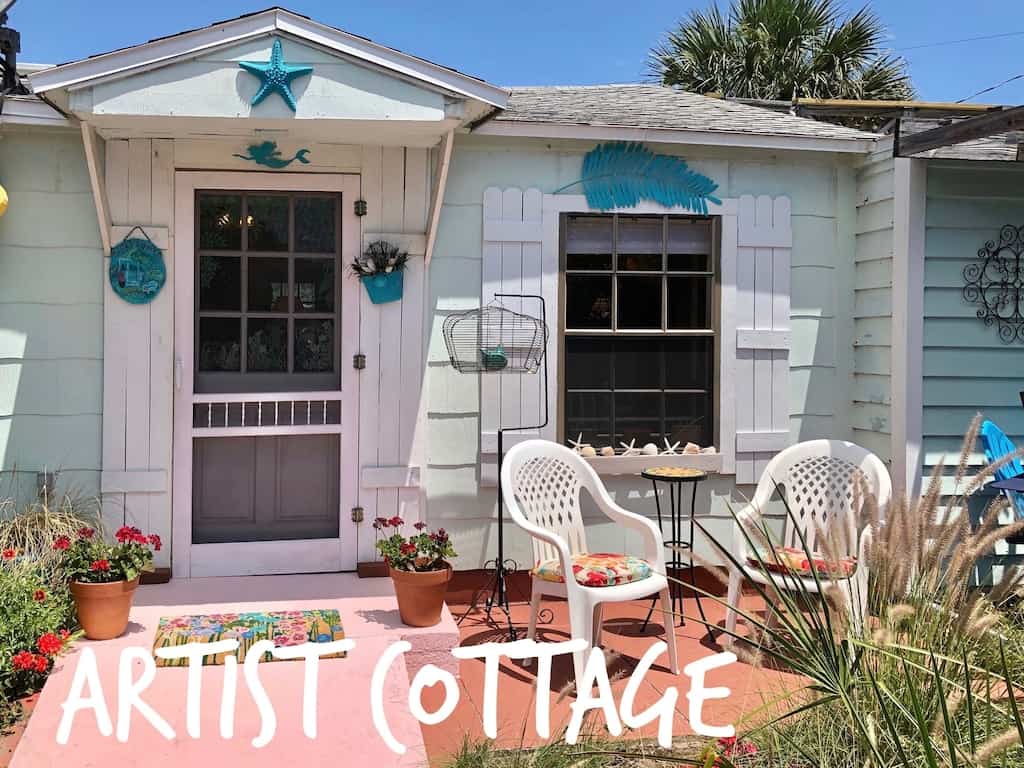 The outside of the turquoise and white artist's cottage. It has a pink pathway and orange porch. 