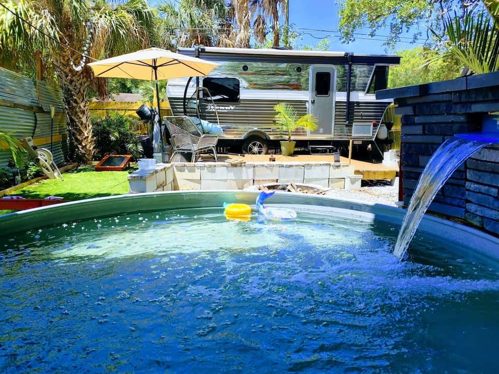 The exterior of the retro trailer. You can see its in a backyard full of palm trees with patio furniture and a small pool. 