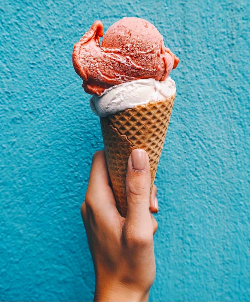 A person with long nails holding an ice cream cone. The cone has a pale white ice cream under a dark pink ice cream. It is being held against a bright blue wall. 