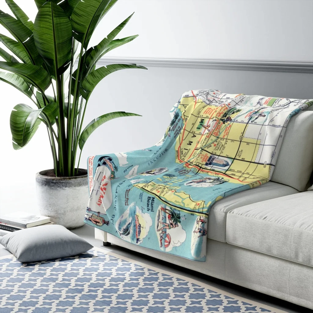 Picture of a fleece blanket with a map of Miami printed on it, draped over a white couch in a generic living room staging featuring some books, rugs, pillows, and a plant