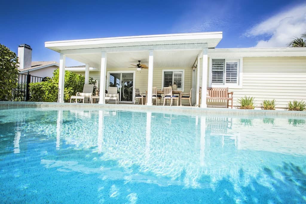 View of a pool and a charming white cottage with a covered patio for lounging and dining. 
