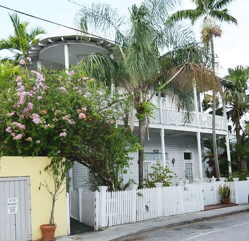 A vintage whitewashed two story house with a wraparound porch framed by palm trees and pink flowers. 