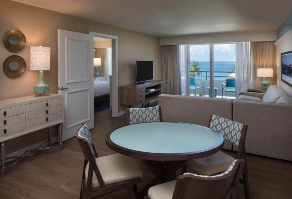 The interior of a beachside suite in a beachfront hotel in Naples Florida. There is a dining table, a large sectional sofa, a separate bedroom, and a private balcony that looks out onto the ocean. 