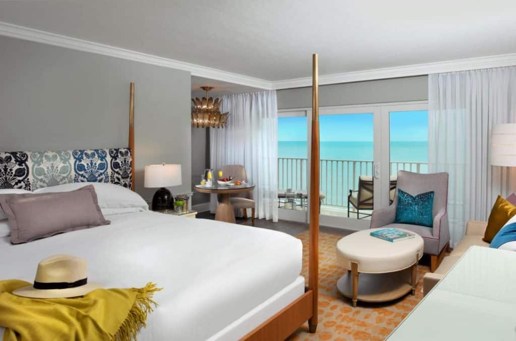 The interior of a luxury hotel on the beach in Naples Florida. It has a king-sized bed, a small bistro table, a chair and ottoman, and a private balcony that looks out onto the ocean. 