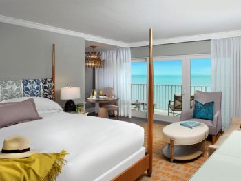 The interior of a luxury hotel on the beach in Naples Florida. It has a king-sized bed, a small bistro table, a chair and ottoman, and a private balcony that looks out onto the ocean.