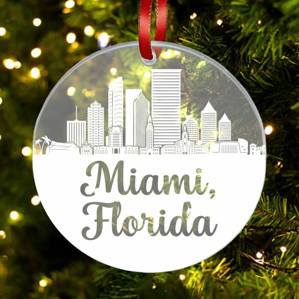 A picture of a glass-like christmas ornament with the Miami Skyline etched into it, on a red bow with a background of christmas trees